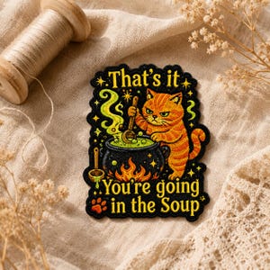Op de afbeelding: Geborduurde patch met een oranje kat die in een borrelende ketel roert met de tekst "That's it, You're going in the Soup." De kat heeft een boze uitdrukking. De patch heeft een zwarte achtergrond met gele sterren en vlammen.