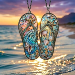 May include: Two pendant necklaces shaped like flip-flops, featuring intricate designs of seashells, seahorses, and starfish. The pendants have a stained-glass effect with blue, teal, and gold colors. The background shows a beach scene at sunset.