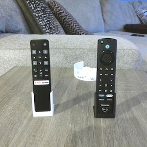 Op de afbeelding: Twee zwarte afstandsbedieningen voor de televisie die rechtop in individuele houders staan. De linker afstandsbediening heeft een Google Assistant-knop en Netflix- en YouTube-logo's. De rechter afstandsbediening is gelabeld met Toshiba en Fire TV.