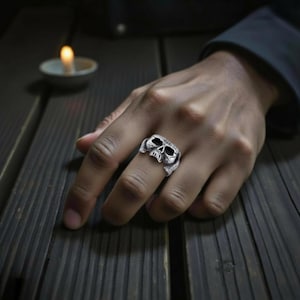 May include: A silver skull ring with detailed facial features, including eye sockets and a jaw. The ring is worn on a finger, showcasing its gothic design. The ring is made of a metallic material and is a piece of jewelry.