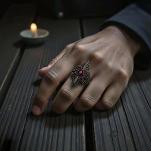 Könnte beinhalten: Ein silberfarbener Ring mit einem detaillierten, verzierten Design, das einen zentralen roten Edelstein und kleinere rote Akzente aufweist. Der Ring wird auf einer Hand vor einem dunklen Holzgrund gezeigt. Eine brennende Kerze ist sichtbar.