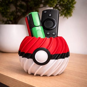 May include: A red, black, and white container in the shape of a Poké Ball, containing a black remote control and two highlighters. The container has a ridged design and is placed on a wooden surface. A small plant is visible in the background.