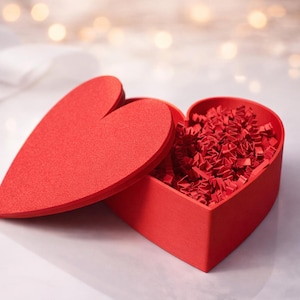 May include: A red heart-shaped box, lid ajar, filled with red paper. The box sits on a white surface, with soft, blurred lights in the background. Ideal for gifts or decorations, the box is a romantic symbol.