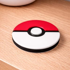 May include: A circular coaster with a red and white design, a black border, and a white button in the centre. It rests on a wooden surface. The design is inspired by a popular animated series.