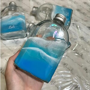 Peut inclure: Une bouteille en verre transparent avec un bouchon à vis argenté, présentant un motif en résine bleu et blanc sur l'extérieur imitant les vagues de l'océan. Le mot Bali est imprimé en écriture blanche sur le verre. La bouteille est rectangulaire.