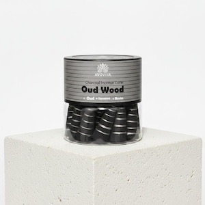 May include: A clear jar of black charcoal incense cones with silver stripes, labeled "Oud Wood." The jar has a silver and gray label with the brand name "AVOVIVA." The jar sits on a white, textured cube.
