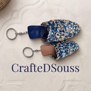 Peut inclure: Deux porte-clés avec des housses crochetées texturées bleu, blanc et beige attachées à des anneaux métalliques. Un porte-clés a une languette en cuir bleu foncé, l'autre une languette beige. Le texte CraftedDSouss est imprimé en dessous.