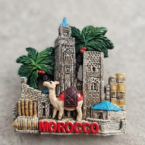 Peut inclure: Un aimant souvenir décoratif représentant en relief une architecture marocaine avec une haute tour, des murs en pierre et des palmiers. Un chameau sellé se tient au premier plan et le mot MOROCCO est inscrit en lettres rouges.
