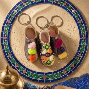 Peut inclure: Trois porte-clés miniatures en cuir de babouche avec des pompons colorés et des broderies complexes. Les porte-clés sont présentés sur une assiette décorative avec une bordure en mosaïque bleue et verte. Une étiquette en bois indique "Crafted D Souss".