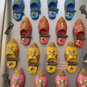 Op de afbeelding: Een verzameling kleurrijke, decoratieve magneten in de vorm van slippers in blauw, rood, geel en roze. Elke slipper heeft een goudkleurige glittertop en sierlijke details. Sommige slippers hebben Arabische tekst. Een boodschappenlijstje is zichtbaar.
