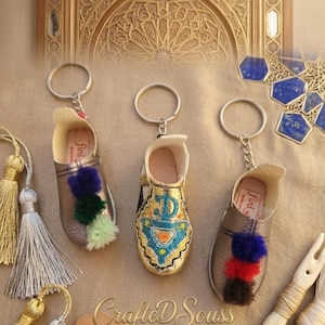 Peut inclure: Trois porte-clés miniatures en forme de babouches en cuir doré et argenté, chacun orné de pompons colorés. Un porte-clés présente des broderies complexes. Les porte-clés sont présentés avec des glands et le texte "Crafted Souss".