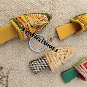 Peut inclure: Quatre porte-clés faits à la main avec des dessus tissés colorés et des bases en cuir ou en bois. Les porte-clés présentent du jaune, de l'orange, du vert et du rose. Le texte "Craftedsouss" est visible sur l'image.