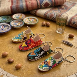 Peut inclure: Quatre porte-clés miniatures en forme de chaussures brodées et colorées sont présentés sur une surface texturée brun clair. Un porte-clés est noir avec "AGADIR" en blanc. Autres éléments : petites assiettes en céramique, noix et un verre.