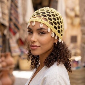 Peut inclure: Un bonnet en crochet jaune pâle avec un motif en maille en forme de losanges. Le bord inférieur du bonnet est orné d'une série de petits coquillages cauris blancs de forme ovale suspendus aux mailles sur tout le pourtour de l'accessoire.