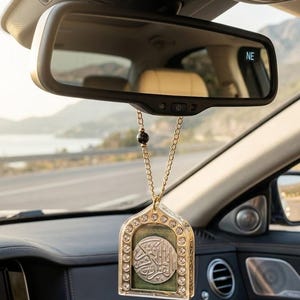 Puede incluir: Un adorno dorado para coche con un diseño decorativo, colgado del espejo retrovisor. El adorno presenta un medallón central plateado con escritura árabe, rodeado de cristales transparentes. El fondo muestra una vista panorámica de una carretera y montañas.