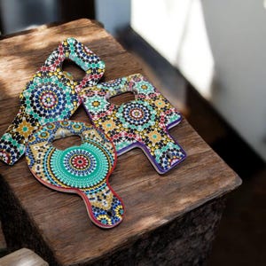 Peut inclure: Trois ouvre-bouteilles décoratifs en bois avec des motifs de mosaïque colorés. Les ouvre-bouteilles présentent une variété de formes, dont un rond et deux avec une poignée. Les motifs de mosaïque incorporent des teintes turquoise, rouges, jaunes et bleues.
