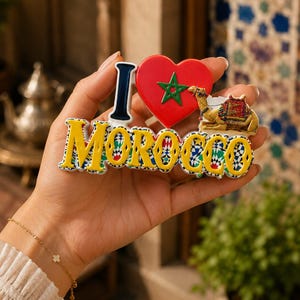 May include: A decorative souvenir magnet featuring the text I LOVE MOROCCO. The letter I is dark blue, the heart is red with a green star, and the word MOROCCO is in yellow with a blue, white, and black geometric tile pattern background.