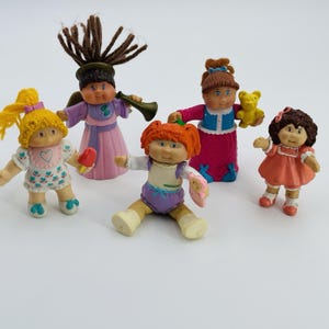 Puede incluir: Una colección de figuras vintage de Cabbage Patch Kids. Las muñecas tienen varios colores de pelo y atuendos, incluyendo vestidos, peleles y ropa temática de ángeles. Una muñeca sostiene una trompeta y otra un osito de peluche.