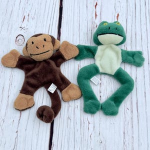 May include: Two plush toys: a brown monkey with tan accents and a green frog with a cream-colored belly. Both toys are lying flat on a light-colored wooden surface. The monkey has a long tail, and the frog has outstretched arms and legs.