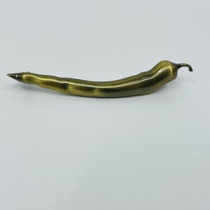 May include: A brass-colored bottle opener shaped like a chili pepper. The opener has a pointed tip and a curved body with a small stem. The metal has a slightly aged, antique appearance.