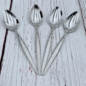 Puede incluir: Cuatro cucharas plateadas con un acabado pulido y reflectante. Los cuencos de las cucharas son de forma ovalada y los mangos tienen un diseño lineal texturizado. Las cucharas están dispuestas sobre una superficie de madera clara.