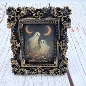 May include: A rectangular picture frame with an ornate, black and gold-brushed finish. The frame holds an illustration of two white ghosts floating above a field of colorful flowers under two crescent moons against a dark, starry night sky.