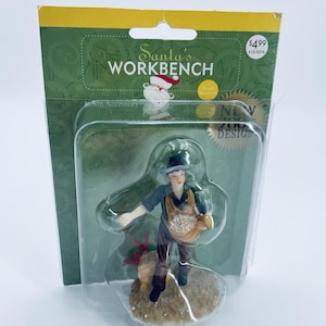 May include: A resin accessory figurine from Santa's Workbench. The figurine depicts a person in a hat and apron holding a basket of white objects. The figurine is on a small, round base. The packaging includes the text "Santa's Workbench" and "New Design."