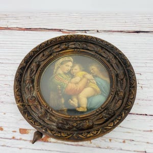 May include: A vintage, oval-shaped, framed print featuring a religious scene. The print depicts three figures in colorful robes, with a central figure holding a child. The frame is made of dark wood with ornate carvings and a small stand.