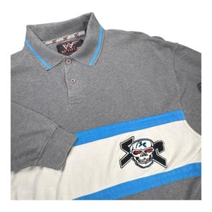 Könnte beinhalten: Ein graues Poloshirt mit einem Kragen mit hellblauer Zierleiste, einer Knopfleiste mit drei Knöpfen und einem WWE Attitude-Etikett. Der Brustbereich zeigt einen breiten weißen Querstreifen mit hellblauer Umrandung und ein Totenkopfmotiv.