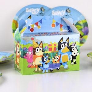 May include: A colorful party favor box featuring cartoon dogs, with the words "Happy Birthday" on the top. The box has a handle and is decorated with images of the characters and a blue sky background. Matching plates are visible in the background.
