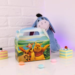 May include: A colorful party favor box featuring Winnie the Pooh and Tigger. The box has a handle and is decorated with a landscape scene. A plush Eeyore toy and small desserts are in the background.