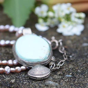 May include: A necklace featuring a light blue pendant and a strand of pink pearls. The pendant is round, set in silver, and has a small seashell charm. The pearls are small and closely spaced, creating a delicate appearance.