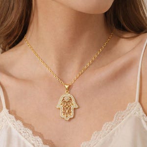 May include: Gold-coloured necklace with a Hamsa hand pendant. The pendant has a detailed filigree design and is embellished with small, sparkling crystals. The necklace has a fine chain and is worn around the neck.