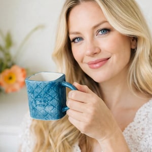 May include: A blue ceramic mug with a quilted pattern. The mug has a square shape and a handle. The interior of the mug is white. The mug is held by a person.