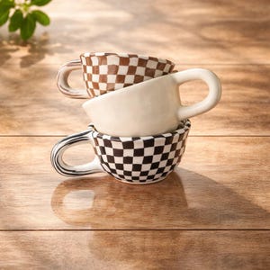 Puede incluir: Tres tazas de té de cerámica apiladas. La taza inferior tiene un patrón de cuadros blancos y negros. La taza del medio es de color blanco liso. La taza superior tiene un patrón de cuadros marrones y blancos. Todas las tazas tienen un asa curva.