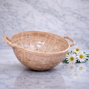 May include: A beige ceramic colander with two handles. The bowl has numerous small holes and a textured surface. The colander is displayed on a marble surface, with white daisies with yellow centres in the background.
