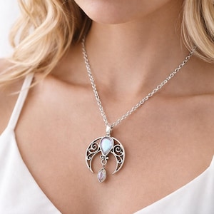 Puede incluir: Collar plateado con un colgante de media luna, con una piedra iridiscente en forma de pera y una piedra más pequeña en forma de lágrima. El collar se lleva sobre una camiseta blanca.