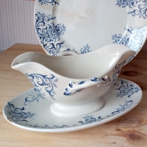 Puede incluir: Un juego de vajilla vintage de porcelana crema, que incluye una salsera, un plato ovalado y un plato grande. Todas las piezas presentan un diseño floral y de volutas en azul. El conjunto se exhibe sobre una superficie de madera.
