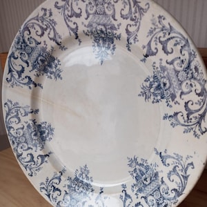 Puede incluir: Un plato de cerámica blanca con un diseño floral y de volutas azules. El plato presenta un espacio central en blanco rodeado por un borde detallado de flores y follaje estilizados. El plato se exhibe sobre un soporte metálico.