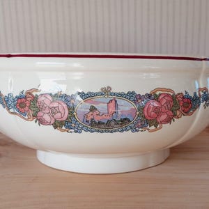 Puede incluir: Cuenco de cerámica color crema con un diseño floral y paisajístico decorativo. El cuenco presenta un borde granate y una imagen ovalada central de un castillo. Rosas rosas y detalles florales azules adornan el exterior.