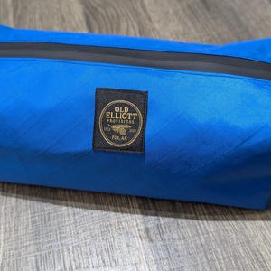 May include: A bright blue rectangular bag with a black zipper and black handles. A black label with gold text reads "OLD ELLIOTT PROVISIONS FOX, AK". The bag is made of a textured material.