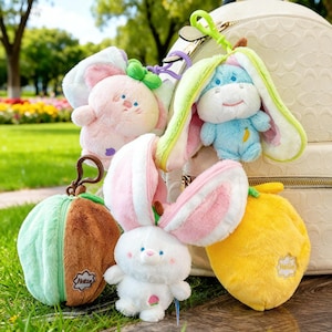May include: A collection of plush bunny and fruit-shaped keychains in various colors. The plushies include a pink bunny, a blue hippo, a white bunny, a green and brown watermelon, and a yellow lemon. Each keychain has a metal clasp for easy attachment.