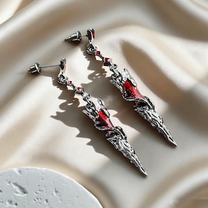 May include: A pair of long, dangling earrings featuring a silver-toned metal finish with intricate, textured detailing. Each earring incorporates vibrant red enamel accents and small, square-cut red stones along the upper chain and central spike.