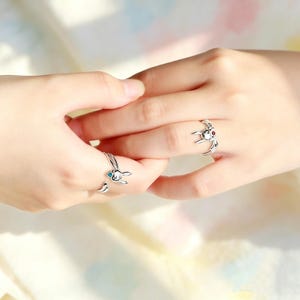 May include: Two silver-colored bunny rings, one with a blue gem and the other with a red gem, are worn on fingers. The rings have a detailed bunny design with curved bands. The background is soft and out of focus.