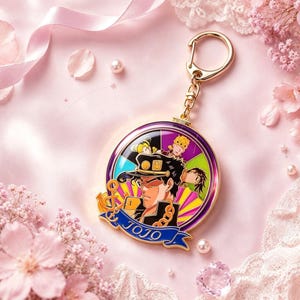 May include: A circular gold-toned metal keyring featuring a colourful enamel illustration of anime characters, including a central figure in a dark cap. A blue banner at the bottom displays the text JOJO in gold lettering, with an anchor icon to the left.