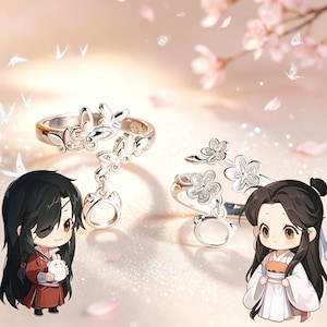 May include: Two silver-toned rings featuring delicate metalwork. One ring displays butterfly motifs with a small dangling circular charm, while the other ring showcases floral designs with a matching dangling circular charm, set against a soft background.