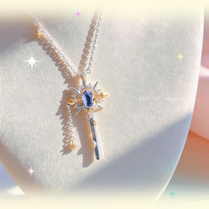 May include: A silver necklace with a pendant shaped like a key. The pendant features a blue gemstone, gold accents, and a starburst design. A small chain with star charms hangs from the key. The necklace is displayed on a white surface.