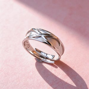 Könnte beinhalten: Ein silberfarbener Metallring mit offenem, verstellbarem Banddesign, das ein poliertes, sich kreuzendes X-Muster über einer matten, strukturierten Oberfläche aufweist. Auf der Innenseite des Rings sind teilweise eingravierte Buchstaben sichtbar.