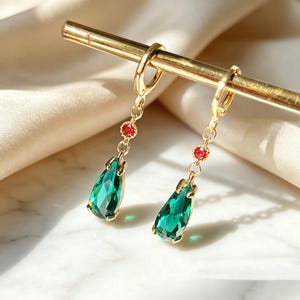 Op de afbeelding: Goudkleurige oorbellen met een druppelvormige, smaragdgroene edelsteen en een kleine rode accentsteen. De oorbellen hangen aan een gouden ring en worden gepresenteerd op een gouden staaf tegen een zachte, crèmekleurige achtergrond.