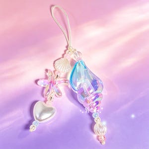 May include: Iridescent seashell and starfish charm with a pearlescent heart and beaded accents. The charm is suspended from a white cord, creating a whimsical, ocean-themed accessory. The charm is set against a gradient pink and purple background.